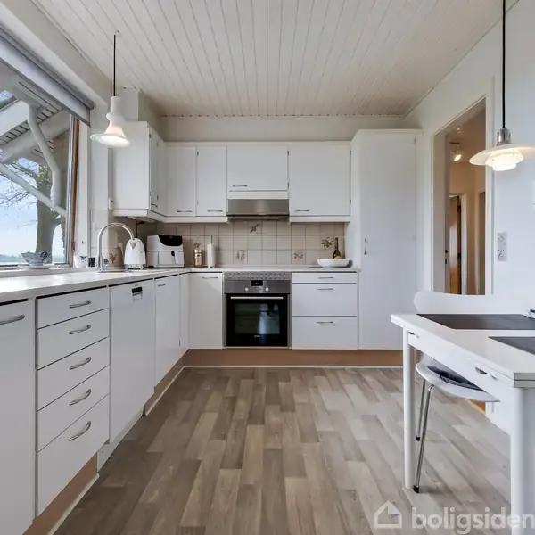 Et lyst køkken med hvide skabe og apparater. En ovn er centralt placeret under en kogeplade. Store vinduer giver udsigt til et træ og himmel udenfor.