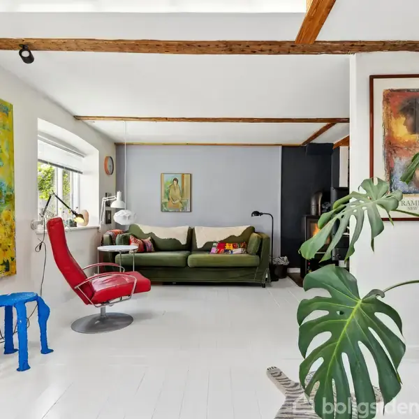 Rød lænestol står ved siden af farverige taburetter i en stue med grøn sofa. Store malerier pryder væggene, og en stor plante tilføjer liv til rummet.