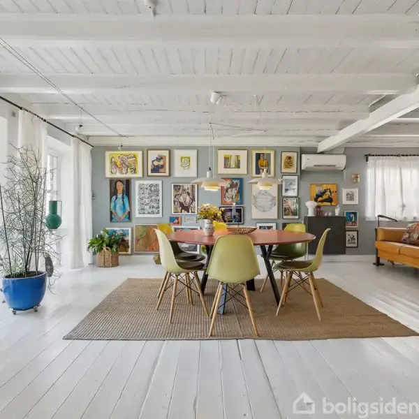 Et rundt spisebord med seks grønne stole står på et tæppe i en stue. Væggene prydes af malerier, og vinduerne har hvide gardiner. En orange sofa står ved siden af en stor bogreol.