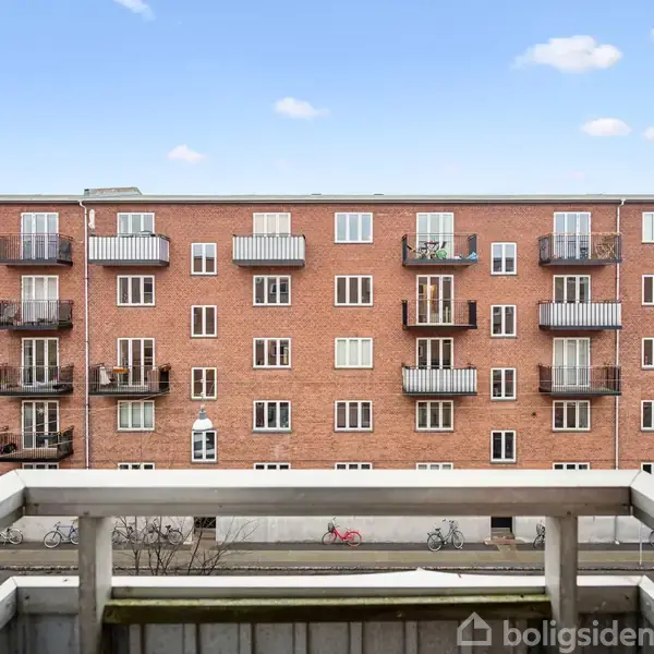 Balkon med hvidt rækværk i forgrunden kigger ud på en murstensbygning med flere altaner. En stille gade med cykler er synlig nedenfor under en lyseblå himmel med få skyer.