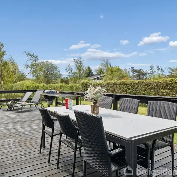 Terrassebord med seks stole står på en træterrasse. I baggrunden er en grill og liggestole. Haven er omkranset af buske og træer under en klar blå himmel.