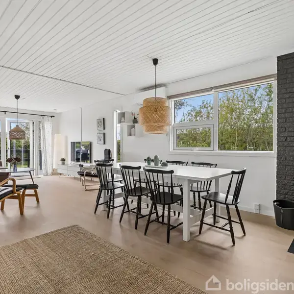 Stue med hvide vægge og loft, indeholder spisebord med stole. Brændeovn til højre og store vinduer med udsigt til have. Tæppe og lænestol til venstre.