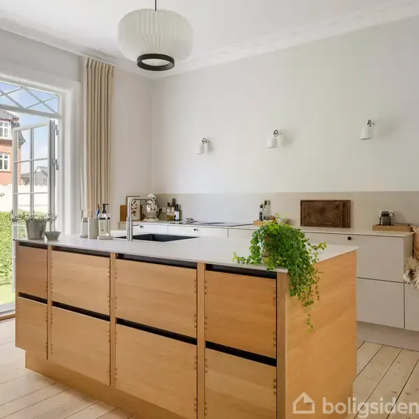 Et lyst køkken med en stor køkkenø i træ, prydet med planter og dekorative genstande. Store vinduer åbner sig til en grøn have udenfor, hvilket giver naturligt lys.