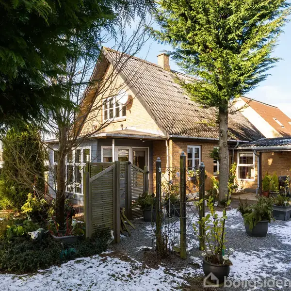 Et brunt murstenshus med skråt tag står omgivet af en have med snepletter og træer. En terrasse med planter i potter ses i forgrunden.