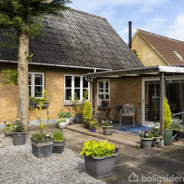 Et gult murstenshus med et skråt tag og en lille, hyggelig terrasse. Terrassen har blomsterkrukker og havemøbler. Træer og buske omkranser gårdspladsen med grusbelægning.