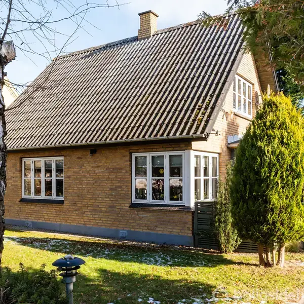 Et gult murstenshus med skråt tag står i en have. Huset har hvide vinduesrammer og er omgivet af træer og græs. Sollys skinner på husets facade.