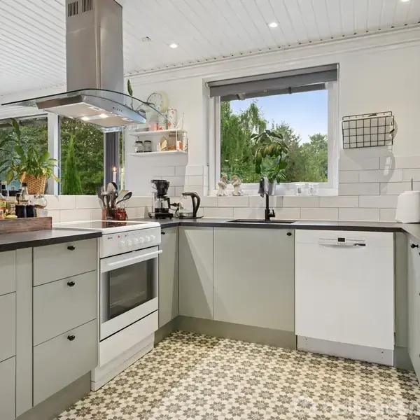 Et moderne køkken er udstyret med hvidevarer, herunder en ovn og opvaskemaskine. Rummet har grønne planter i vindueskarmen og dekorative fliser på gulvet, med udsigt til udendørs grøntområde.