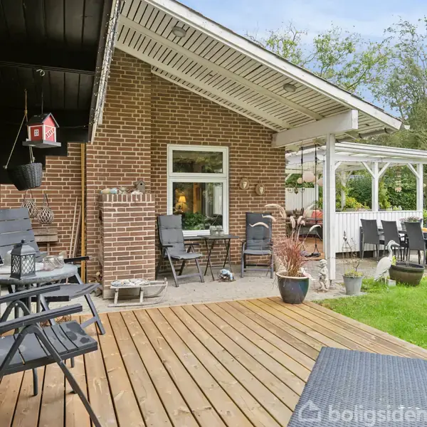 En udendørs træterrasse med sorte havestole står foran et murstenshus. Til højre ses en overdækket siddeplads med bord og stole. Omkring er der græsplæne og buske.