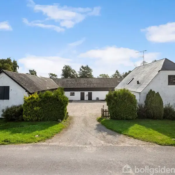 To hvidkalkede bygninger står på hver side af en grusvej, der leder til en større bygning i baggrunden. Omgivet af græsarealer og træer under en blå himmel.