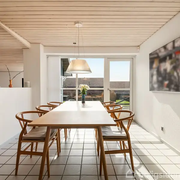 Spisebord med seks stole står på flisegulv. En stor lampe hænger over bordet, og der er en vase med blomster. Bagved ses en glasdør med udsigt og et ophængt billede på væggen.