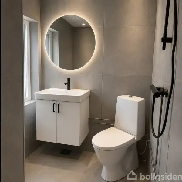 Rundt spejl lyser op over en hvid vask med sort hane. Toilet ved siden af i et moderne badeværelse med grå fliser på væggen. Vandhane og spejl tilføjer elegant præg.