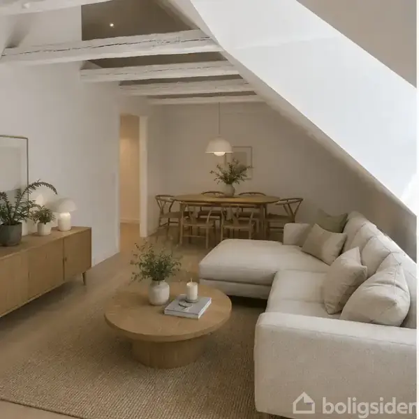 Stue med hjørnesofa i lyse farver, en rund træbord med planter og stearinlys, i et moderne rum med skrå lofter. En spisebord med stole står i baggrunden.