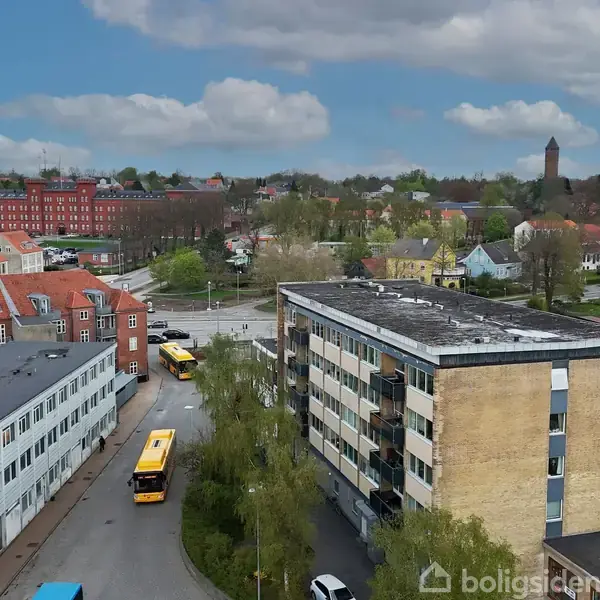 To bygninger i forgrunden med flade tage, omringet af busser og biler på vejene. En række røde teglstenshuse og grønne træområder strækker sig i baggrunden under en blå himmel.