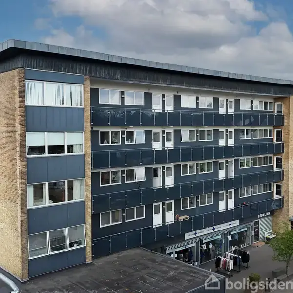 En femetagers bygning med vinduer og mørke facader står i et bymiljø. Foran er der en forretning med tøjstativer. Baggrunden viser andre bygninger og en lys himmel.