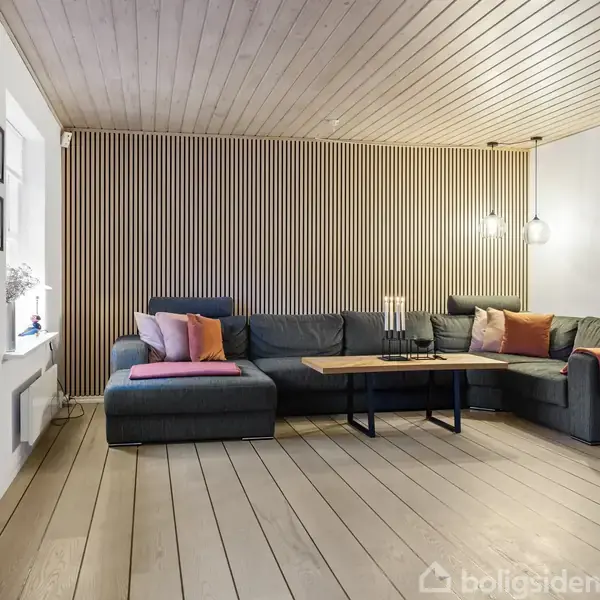 Sofa med farverige puder står i en moderne stue. Den ribbede trævæg og træloft skaber en varm atmosfære. Et sofabord står foran sofaen med levende lys.