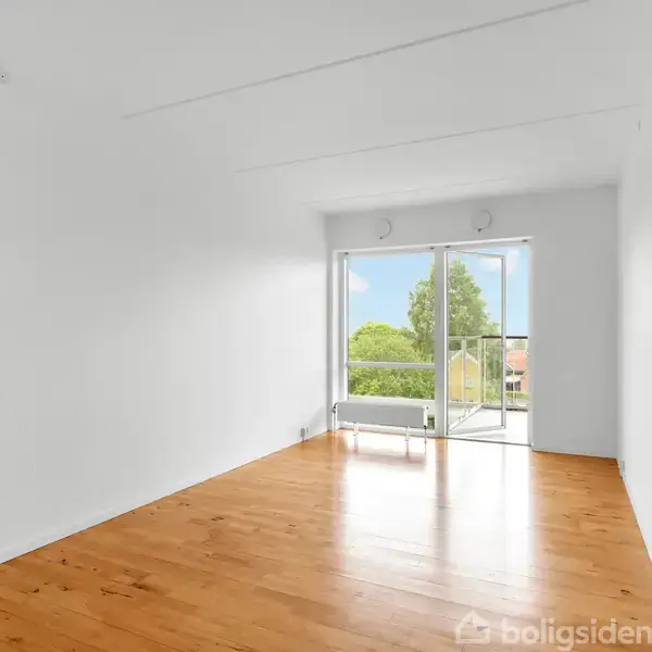 Et tomt, lyst rum med trægulv leder opmærksomheden mod en åben altandør, der fører til en balkon. Udenfor ses grønne træer mod en blå himmel. Ingen tekst til stede.