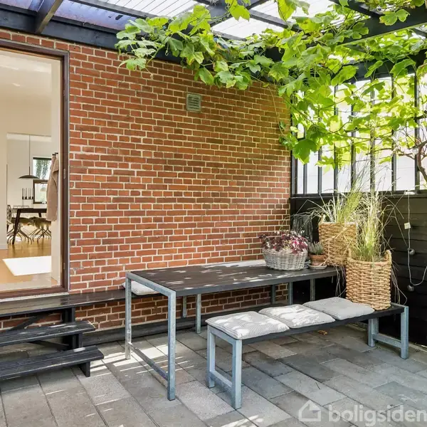 Terrassemøbler står på en flisebelagt terrasse tæt på en murstensvæg. Et bord med to bænke ses under en pergola med planter. En grill placeres til højre for bænken.