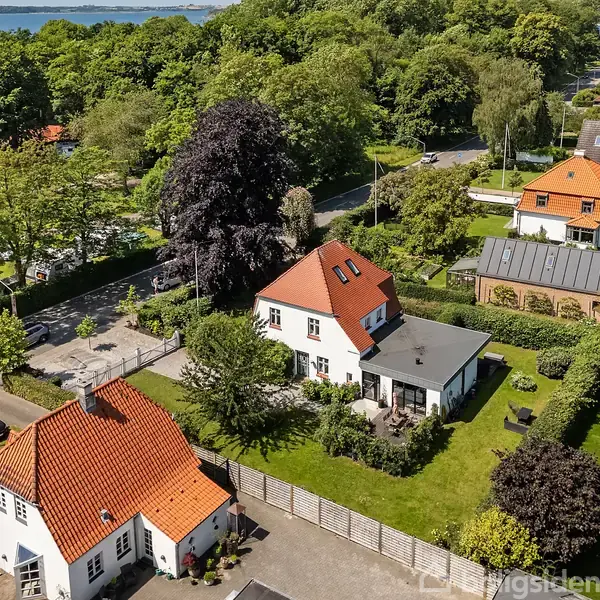 Hus med rødt tag står i en velholdt have med græsplæne. Omgivet af flere huse og grønne områder. En sø skimtes i baggrunden blandt træer.