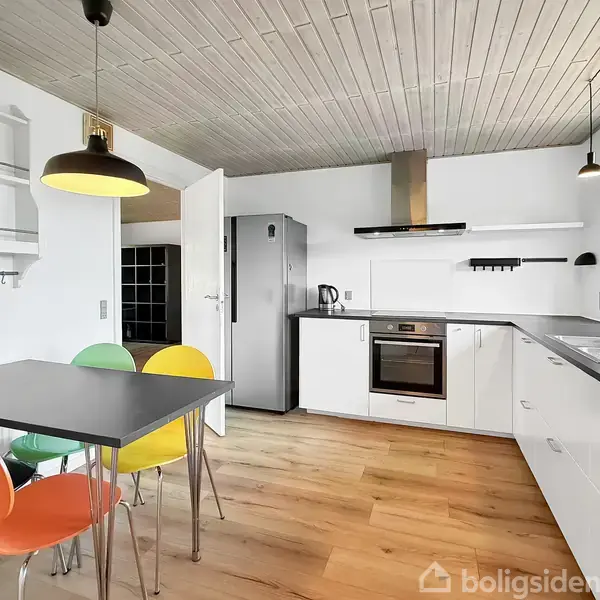 Et moderne køkken med hvide skabe, gråt bord med farverige stole, og indbygget ovn. En stålgrå køleskab står i hjørnet. Trægulve og træloft tilføjer varme. Lampen hænger over bordet.