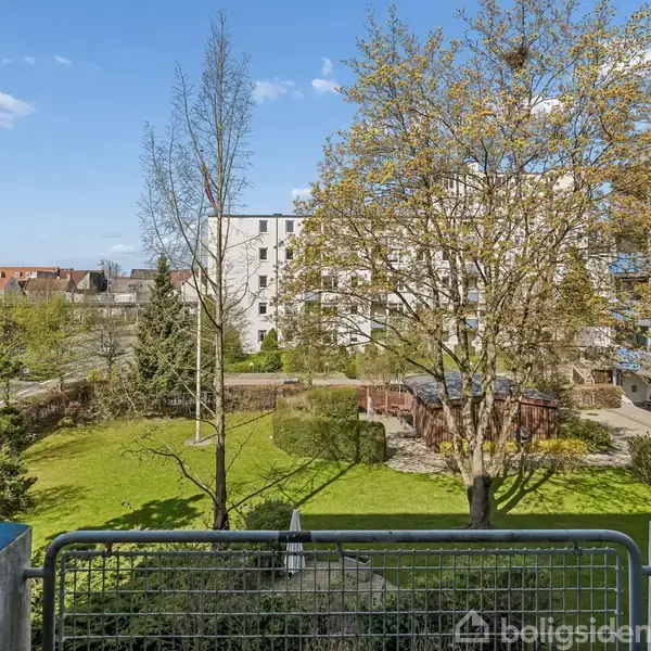Balkon overlooks grøn have med træer og buske. I baggrunden ses moderne lejlighedsbygninger under en blå himmel med få skyer.