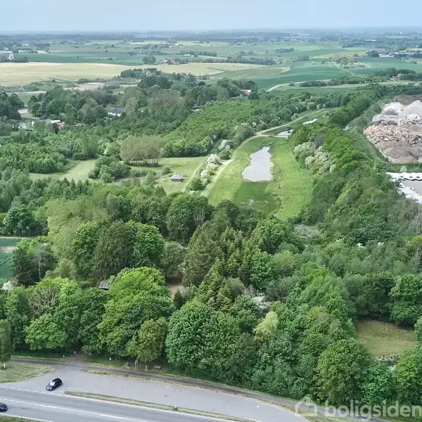 Landskab omgivet af frodig grøn skov med en vej i forgrunden, en smal flod løber gennem området, mens marker og industrielle bygninger står i baggrunden.