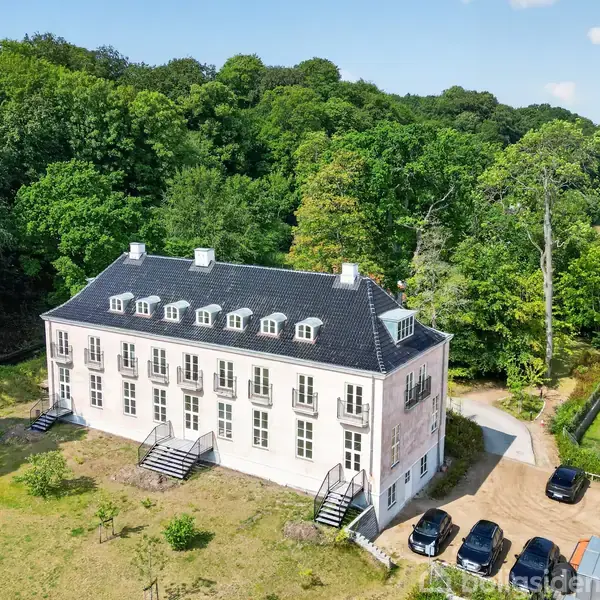 En hvid treetagers bygning med sort tag står omgivet af frodig skov. Den har adskillige små altaner og trapper. Flere biler er parkeret foran på en grusvej.
