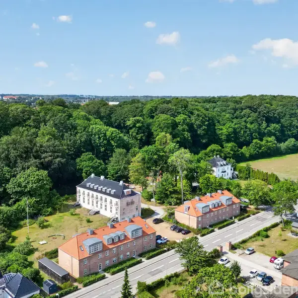 Stort hvidt hus omgivet af frodig skov, til venstre for to mindre, røde bygninger. Huse ligger ved en vej med mindre trafik. Solrig dag med blå himmel og få skyer.