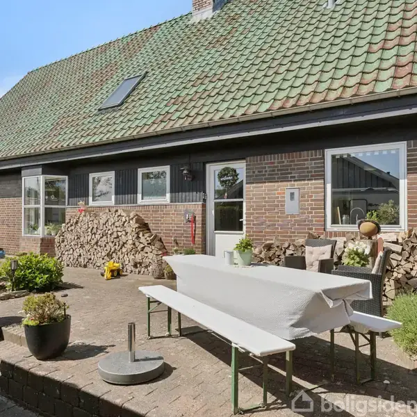 Et langt træhus med grønne tage, omkranset af en gårdhave med blomsterkrukker og en overdækket bænk, ligger på en terrasse belagt med mursten og stablet med brænde langs husmuren.
