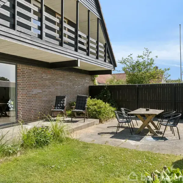 Hus med murstensvæg har en terrasse med sorte stole omkring et bord. En hængestol er til venstre nær en glasdør. Græsplæne og hæk omgiver terrassen.