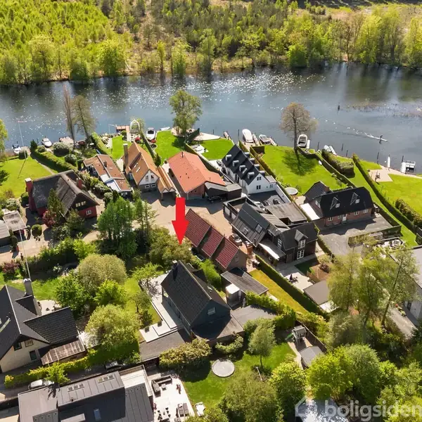 Røde hus, markeret med pil, står blandt andre omkringliggende huse. Det ligger ved flodbredden med frodig vegetation og en bro, omgivet af grønne træer og en lys himmel.