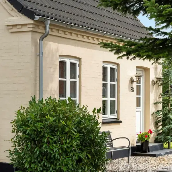 Hus i lysegul mursten med sorte tagsten, har to vinduer og en hvid dør med nummeret "88". En bænk og planter foran, omgivet af grus og træer i baggrunden.