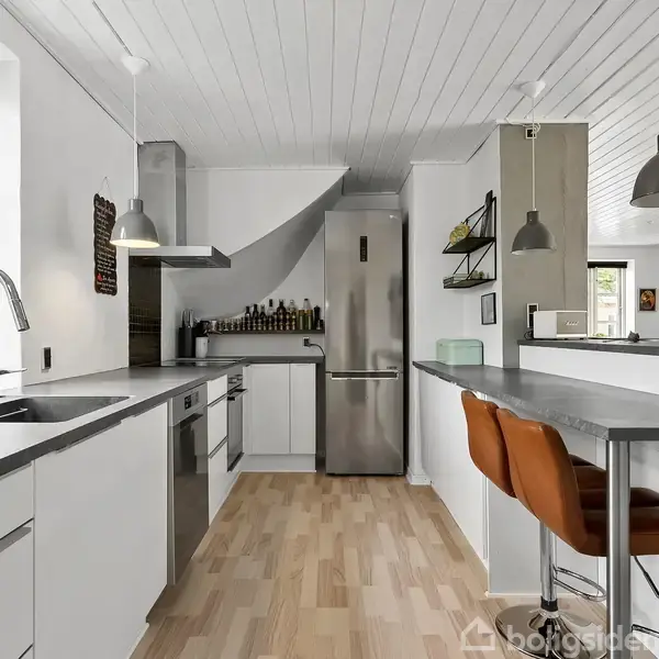 Køkken med moderne indretning; to orange barstole ved køkkenbord, stålvask ved vindue, stålkøleskab, og flere lamper. Trægulv og hvidt loft skaber en lys atmosfære.