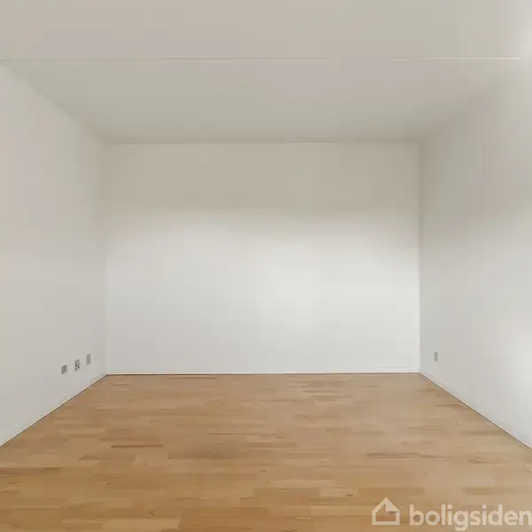 Tomt rum med lyse trægulve og hvide vægge, ingen møbler til stede, hvilket skaber en minimalistisk atmosfære.