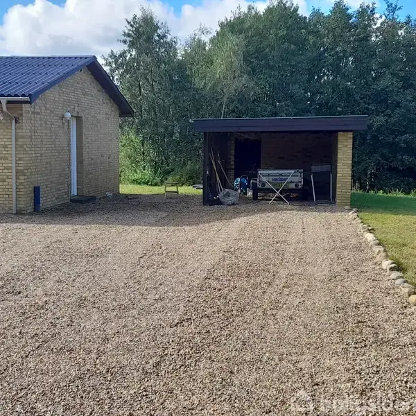 Et lille, gult murstenshus med mørkt tag står til venstre. Til højre ses en carport med forskellige genstande. Omgivelserne er en grusbelagt indkørsel og grønne træer i baggrunden.