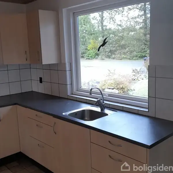 Køkkenvask med hane indbygget i en sort bordplade, omgivet af beige skabe og hvide fliser. Udsigt gennem et vindue viser en have med buske og træer.