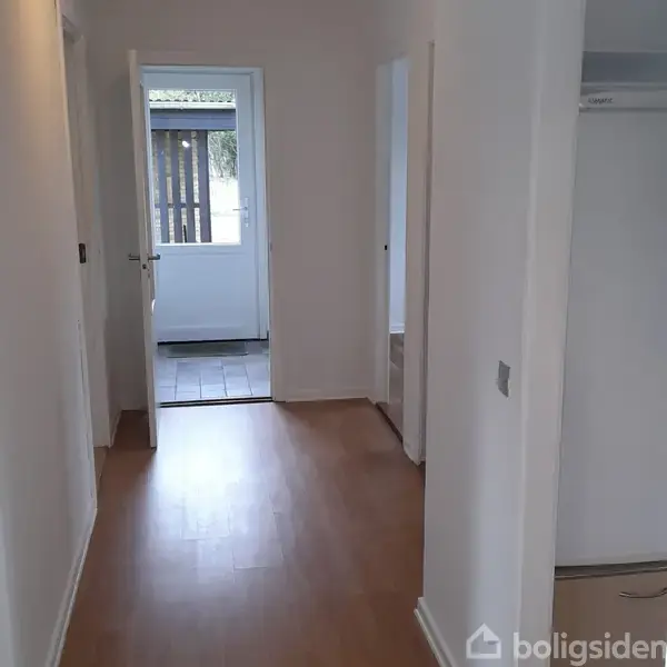 Gang med lys trægulv fører til en dør mod en lysere flisebelagt entré eller terrasse. Væggene er hvide, og der er flere døre på siderne.
