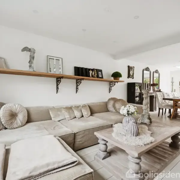 Stor, lys stue med beige sofa og sofabord. Hylde med dekorationer på en hvid væg. Spisestue med langbord og stole nær vinduer i baggrunden.