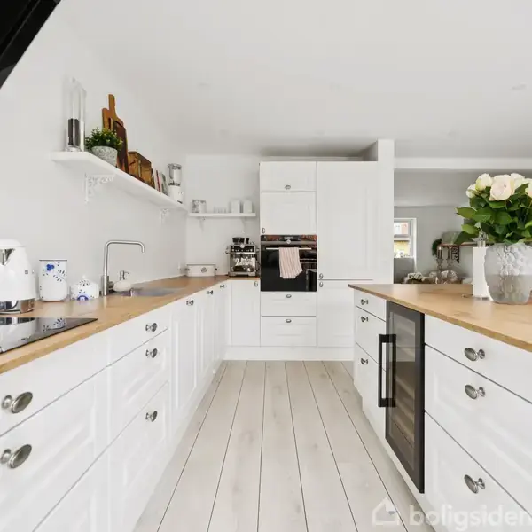 Et lyst, moderne køkken med hvide skabe og træbordplader. En vase med blomster står på en ø. Hylder og køkkenudstyr vises, mens en åben stue ses i baggrunden.