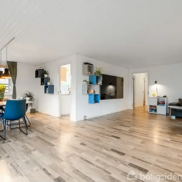 Stue med trægulv; spisebord med stole ved vindue; vægmonteret tv; grå sofa i hjørne; vægklock på hvid væg; hylder med planter.