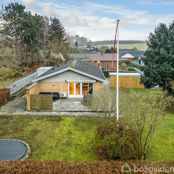 Et enetages hus med gråt tag ligger i en have med grøn plæne. En flagstang står foran, og en trampolin er til venstre. Baggrunden viser flere træer og huse.