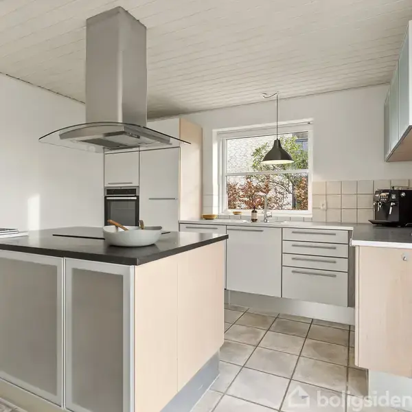 Et moderne køkken med en køkkenø udstyret med en emhætte. Køkkenet har lyse skabe, hvide fliser, et vindue og en kaffemaskine. Flisegulvet og en loftslampe fuldender rummet.