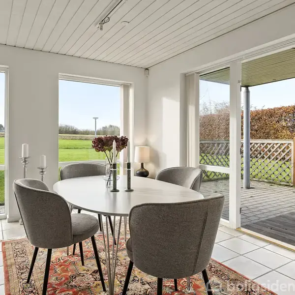 Et ovalt bord med fem grå stole står på et farverigt tæppe. Det er placeret i en lys spisestue med udsigt til en grøn have gennem store vinduer.