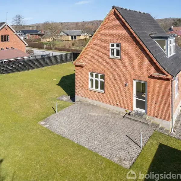 Et rødt murstenshus med sort tag står på en stor, grøn græsplæne. Der er en brolagt indkørsel ved siden af huset i et villakvarter med flere lignende huse.