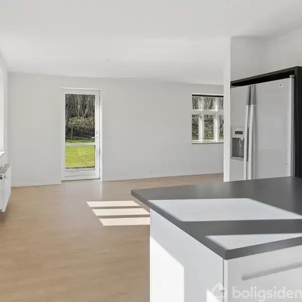 Køkken med hvidt interiør og sort bordplade, en moderne stål-køleskab. Store vinduer og en dør åbner til en have. Et rum belyst af naturligt lys.