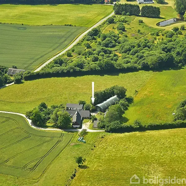 Luftfotografi over en gård omgivet af marker og træer. En hvid pil peger mod bygningen, som er centralt placeret i et grønt, landligt landskab med flere marker og veje.