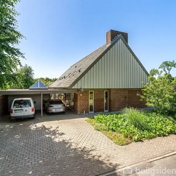 Et rødt murstenshus med et skråt tag står i en have, flankeret af en terrasse. En carport med to biler ved siden af huset. Grønne træer og buske omgiver området.