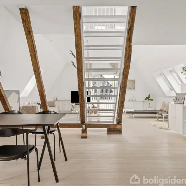 Spisebord med stole står under en sort lampe i en lys loftslejlighed. Synlige træbjælker deler rummet, der har hvide vægge, skråvinduer og moderne indretning med sofagruppe og dekorative planter.