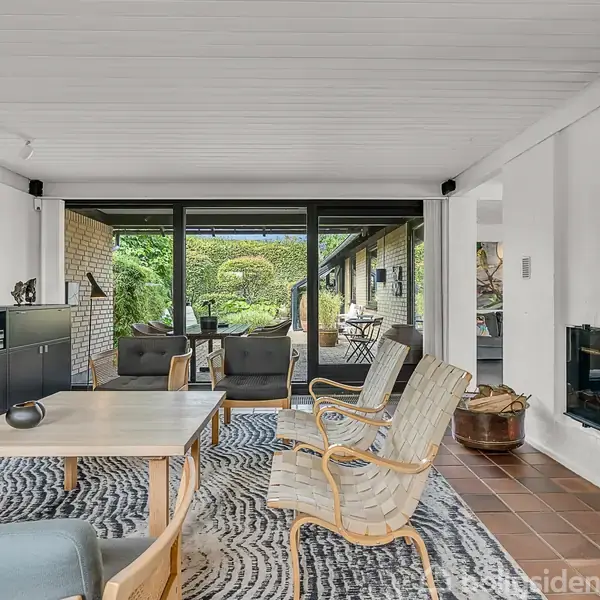 Stue med lyse møbler og stengulv. Store vinduer åbner mod en veranda med udsigt til en have. Fladskærms-TV og kunst på væggene.