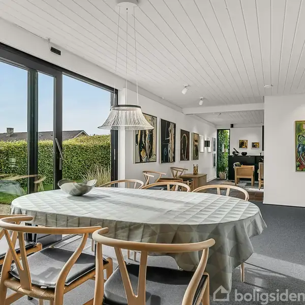 Rundt spisebord med dug står i en spisestue ved store vinduer ud til en have. Væggene er prydet med kunst, og der er et minimalistisk interiør med træstole.