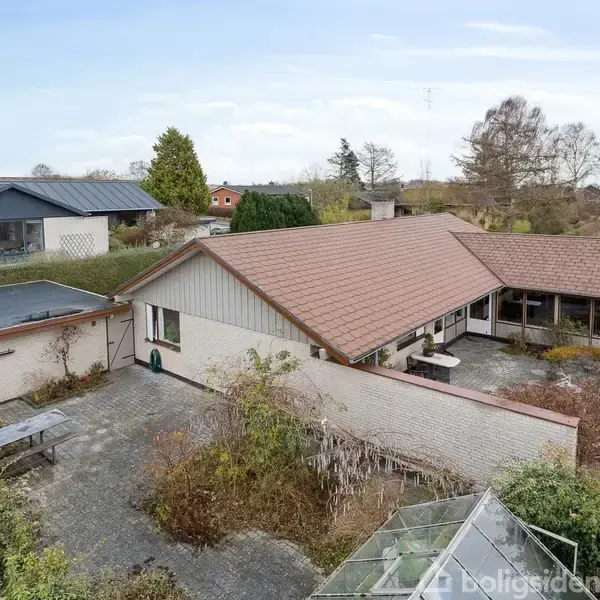 En murstensvilla med rødt tag ses fra fugleperspektiv. Den omkranses af en stor flisebelagt terrasse og have. Andre huse og træer ses i baggrunden under en blå himmel.
